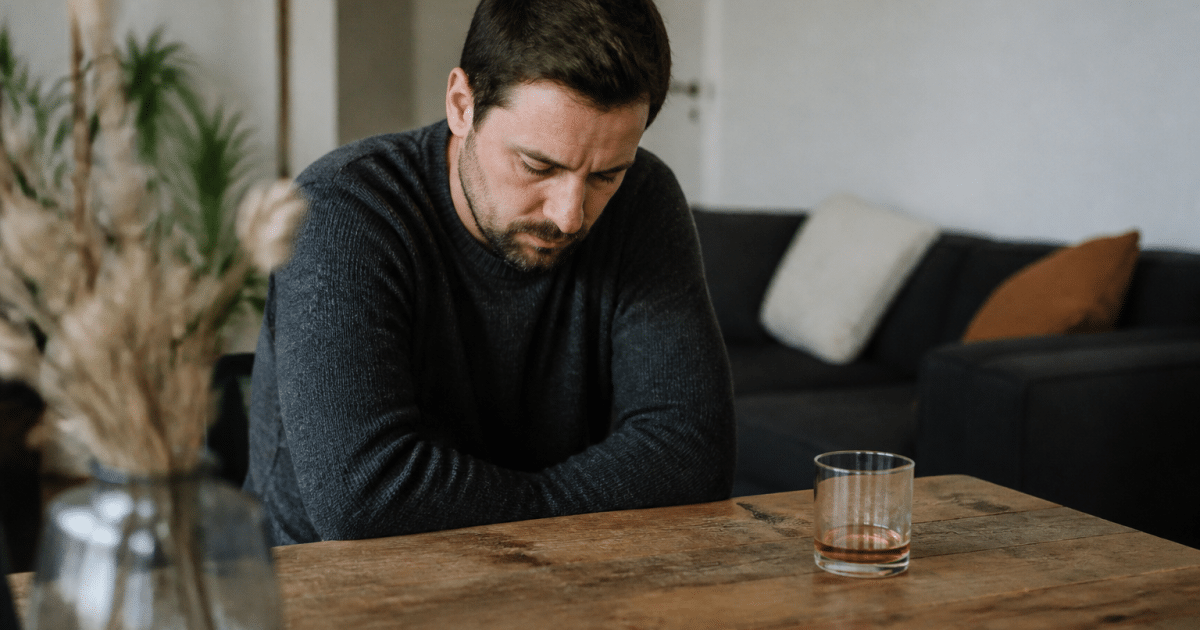Homme assis seul, tête baissée, verre a alcool a proximité.