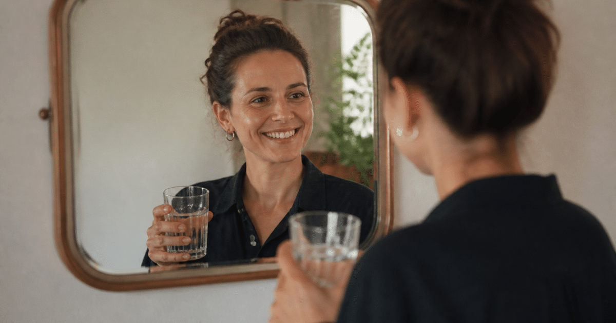 Personne souriante tenant verre eau devant miroir.